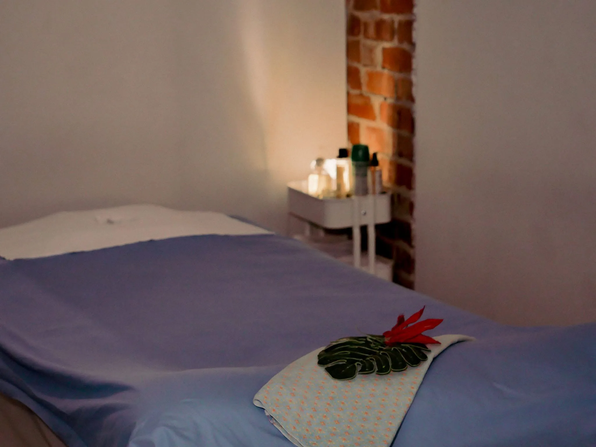 Red hibiscus on folded Thai-print cloth at the foot of a massage bed, warm salt-lamp glow in an exposed-brick nook
