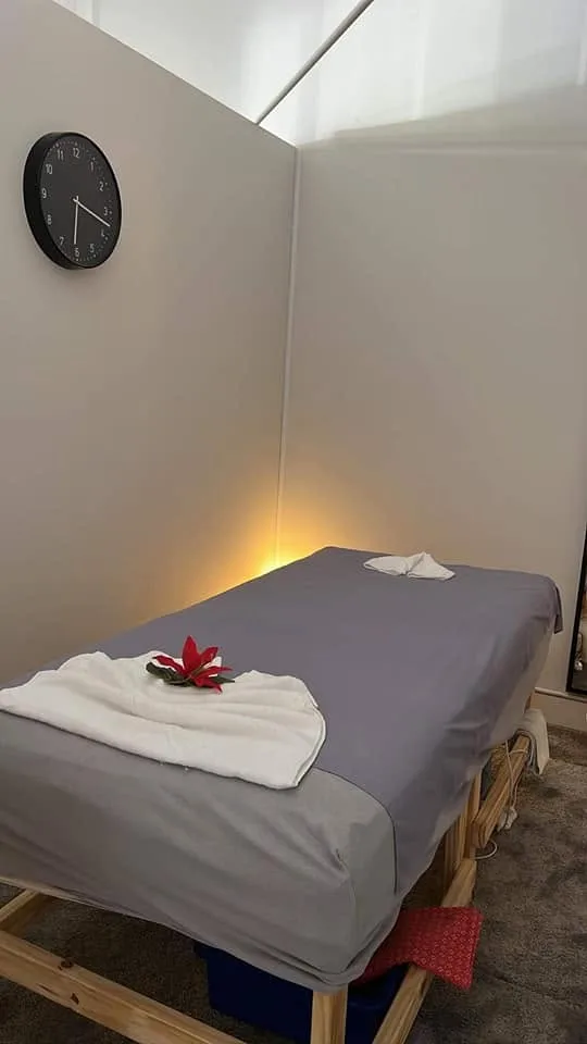 Grey-linen massage bed with folded white towels and a red flower, black wall clock and warm accent light