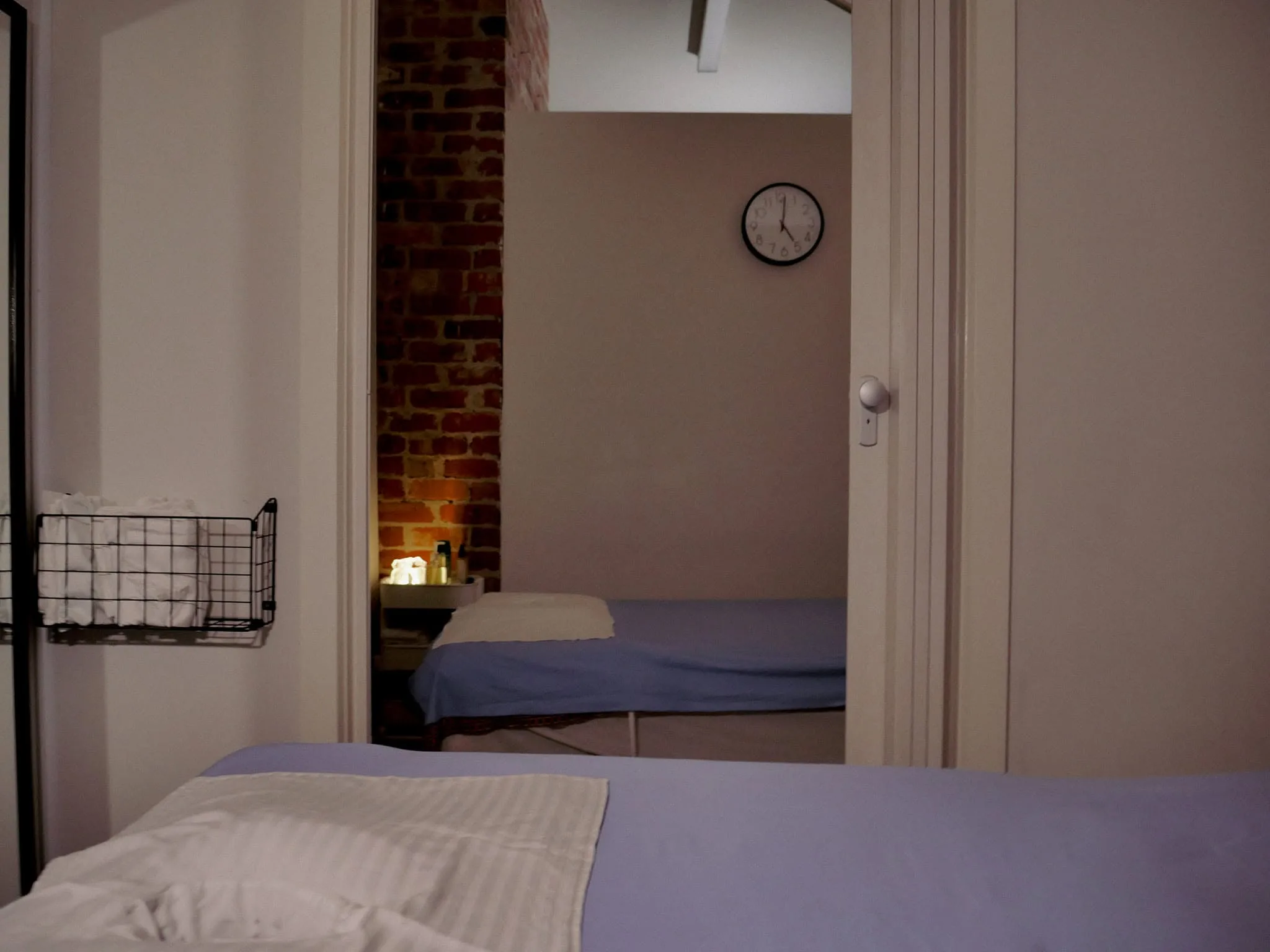 Two treatment rooms at Ladda Thai Massage through a doorway — exposed brick, wall clock and lilac linens