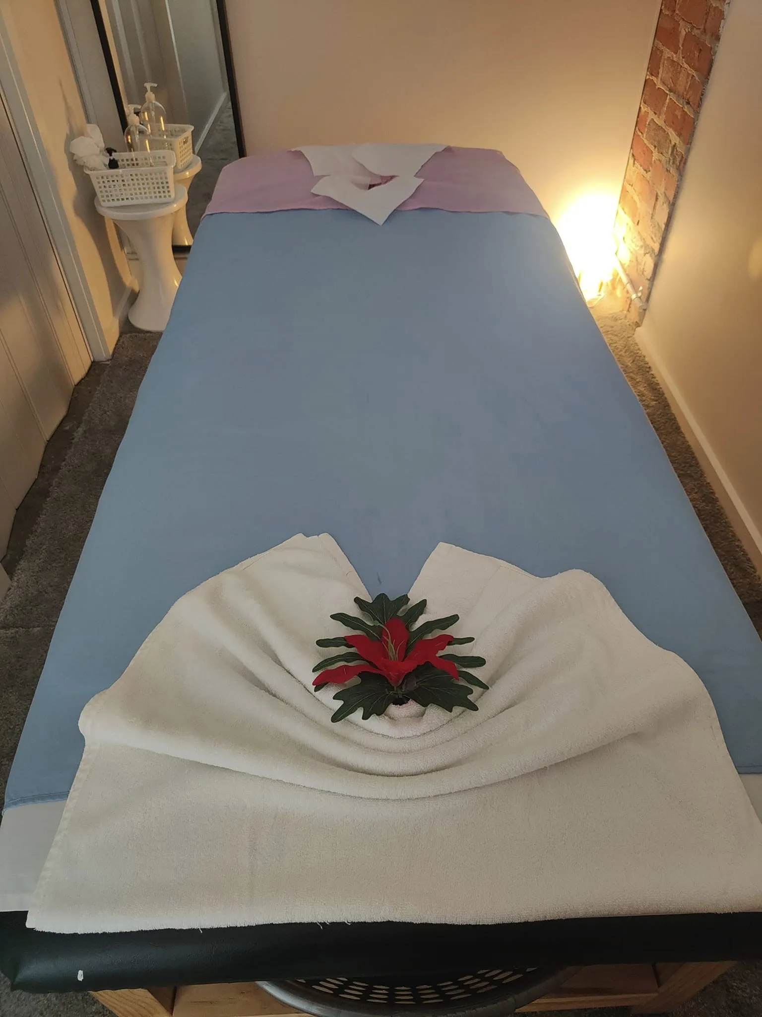 Overhead view of a made massage bed with red hibiscus and lilac linen, exposed brick and warm lamp
